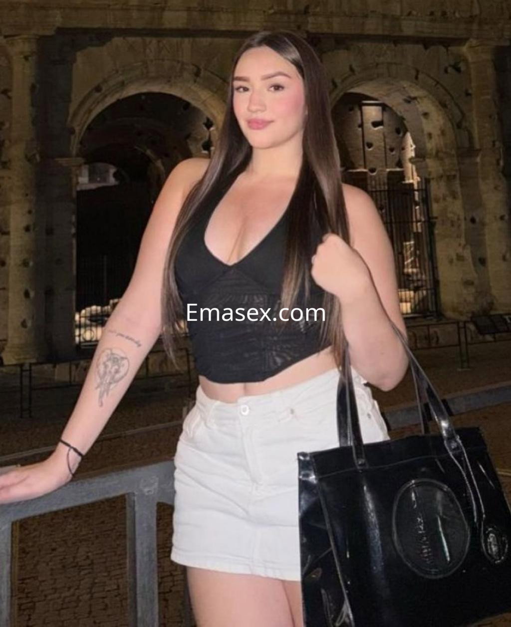 Escort en Barcelona de pie frente a un monumento, vistiendo top negro y falda blanca, con cabello largo y lacio.