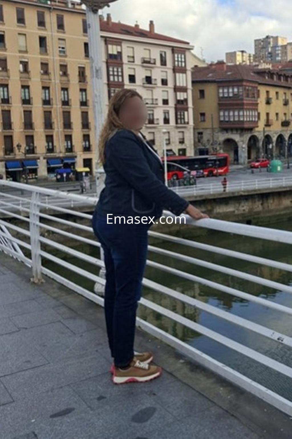 Puta de cabello rizado y rubio, vestida con chaqueta oscura y jeans, apoyada en la barandilla en Bilbao.
