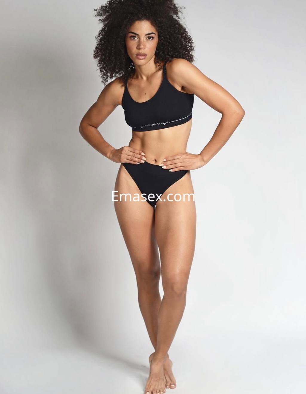 Escort de pie con un top negro y bikini a juego, en un fondo neutro que resalta su figura en Santander.