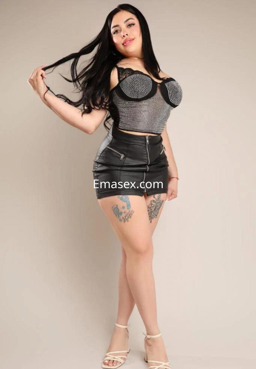 Escort en Toledo con cabello largo y lacio, vistiendo un top de malla y falda corta de cuero, posando con confianza.