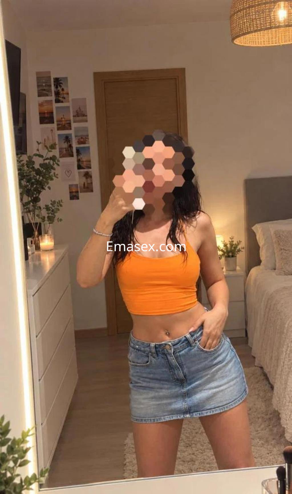 Escort en Tarragona con top naranja y falda de mezclilla, posando frente a un espejo en un ambiente acogedor.