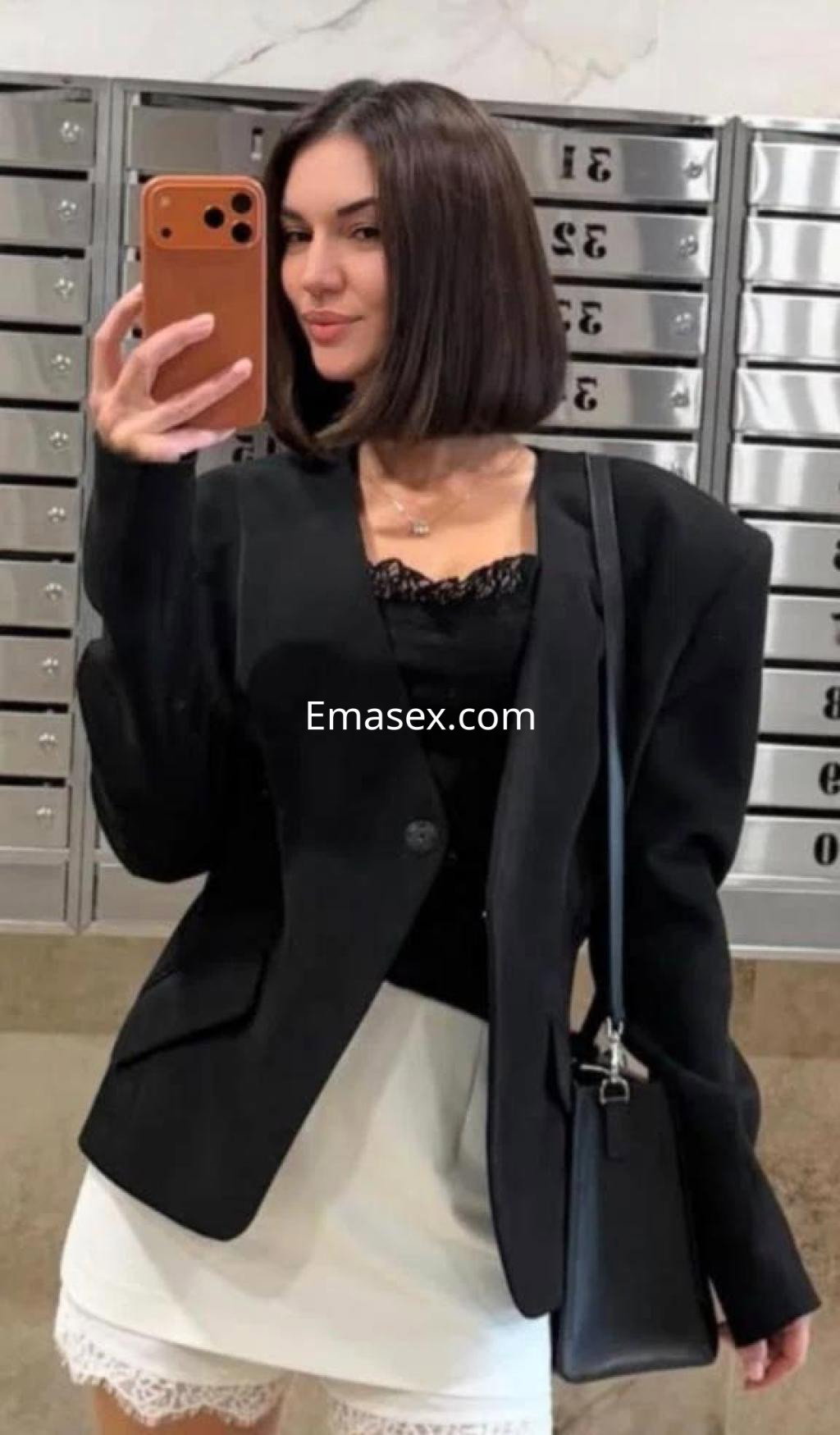 Puta en Cáceres lleva un abrigo negro oversize, blusa negra de encaje y falda blanca, posando en un vestíbulo.