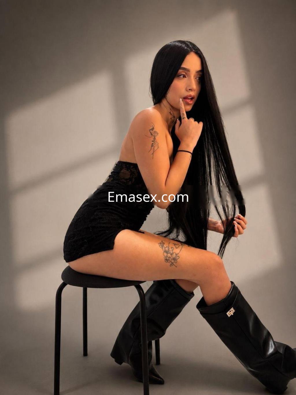 Escort en Girona, sentada sobre una silla, luciendo un vestido negro y botas altas, con cabello lacio y tatuajes visibles.