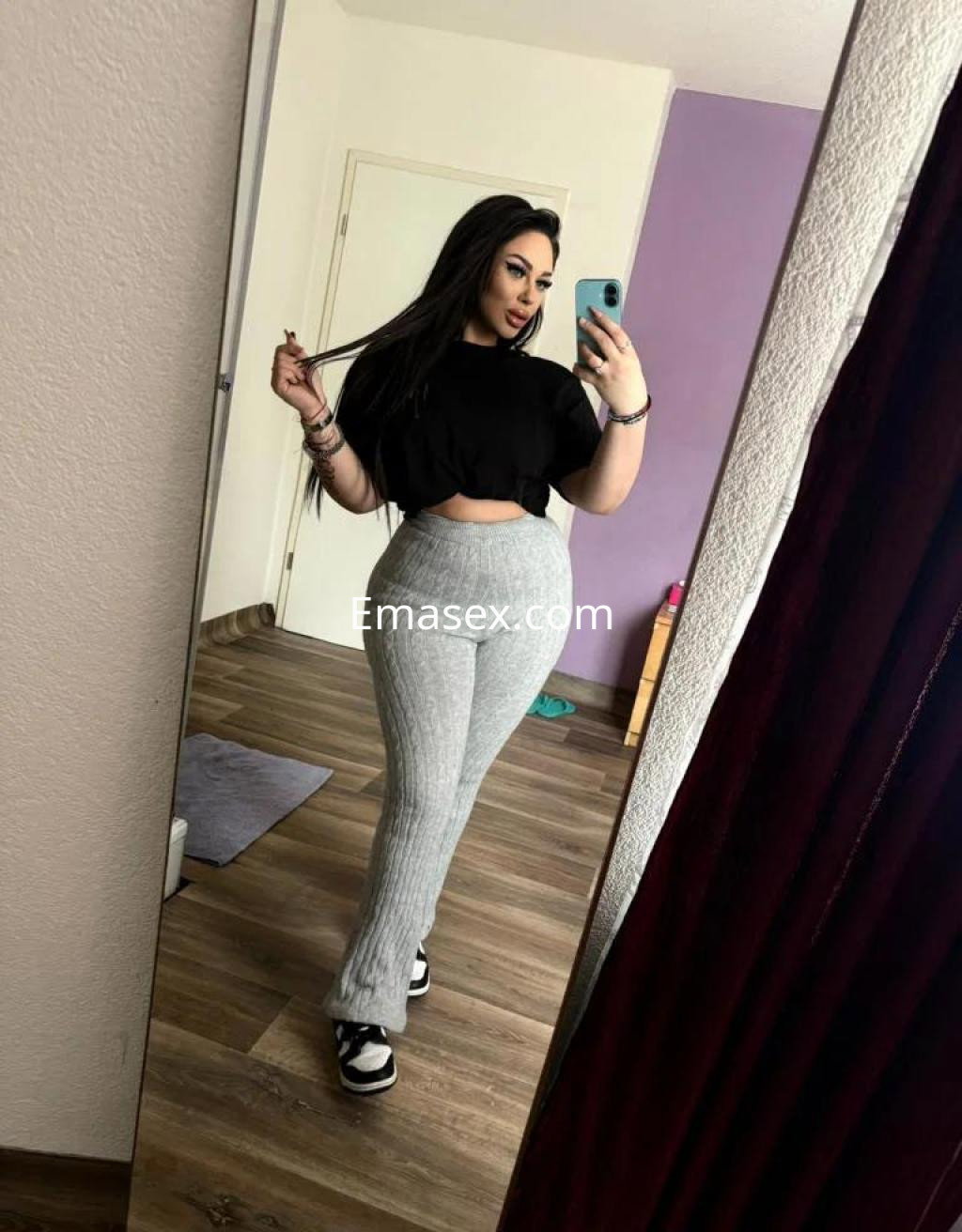 Escort en Girona con cabello largo y oscuro, viste una camiseta negra y pantalones grises ajustados, posando frente al espejo.