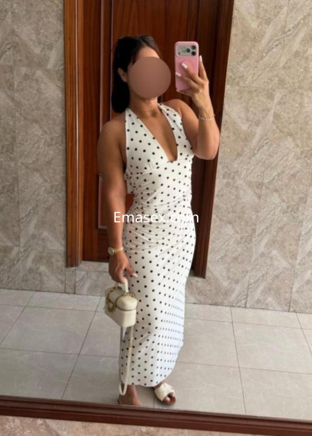 Puta en Castellón posando frente a un espejo, luciendo un vestido blanco con puntos negros y sandalias.