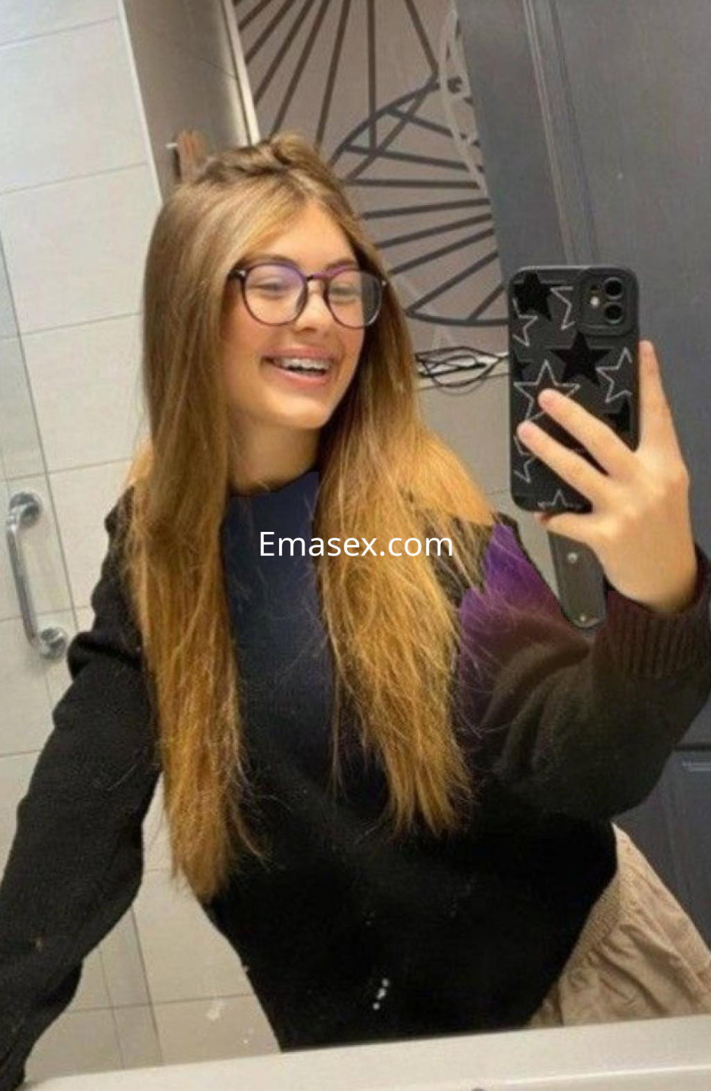 La puta en Córdoba sonríe mientras toma una selfie en el espejo, luciendo un suéter negro y cabello largo.