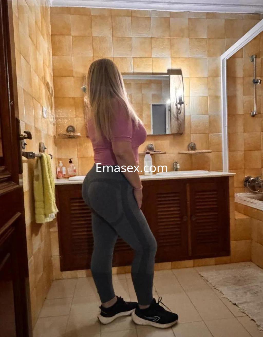 Puta en Huelva con cabello largo y rubio, vestida con camiseta rosa y leggings gris oscuro, posando de perfil en un baño.
