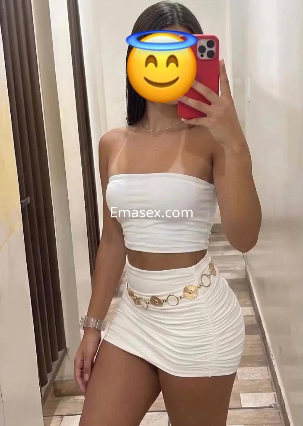 Escort en Las Palmas con blusa blanca sin mangas y falda ajustada, posando frente a un espejo.