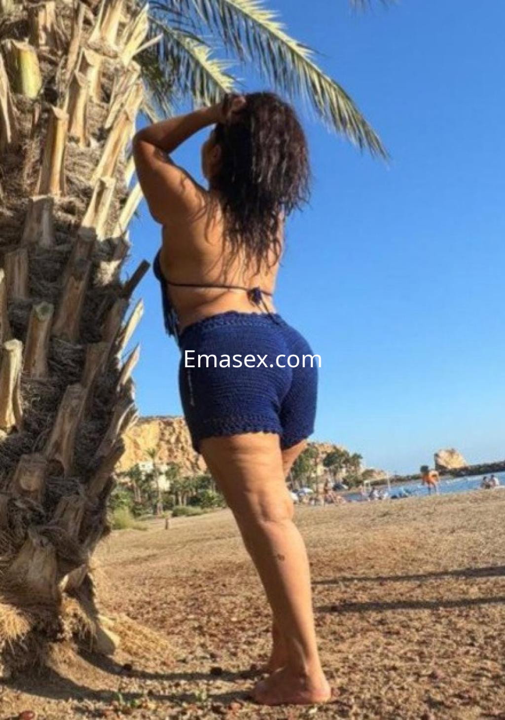 Puta de Albacete mostrando su figura en la playa, con un top azul y shorts cortos, junto a una palmera.
