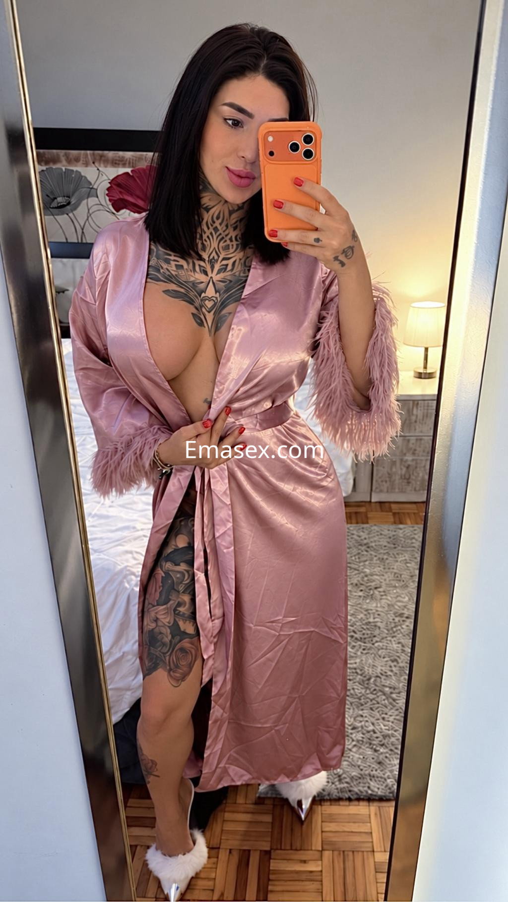 Puta trans en Vigo con bata de seda rosa, mostrando tatuajes y posando frente a un espejo en un ambiente acogedor.