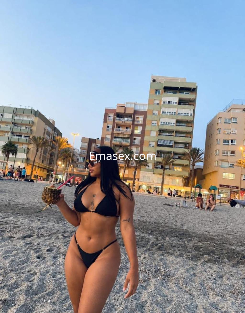 Escort en Almería, luciendo un bikini negro y gafas de sol, sostiene una bebida en un ambiente playero.