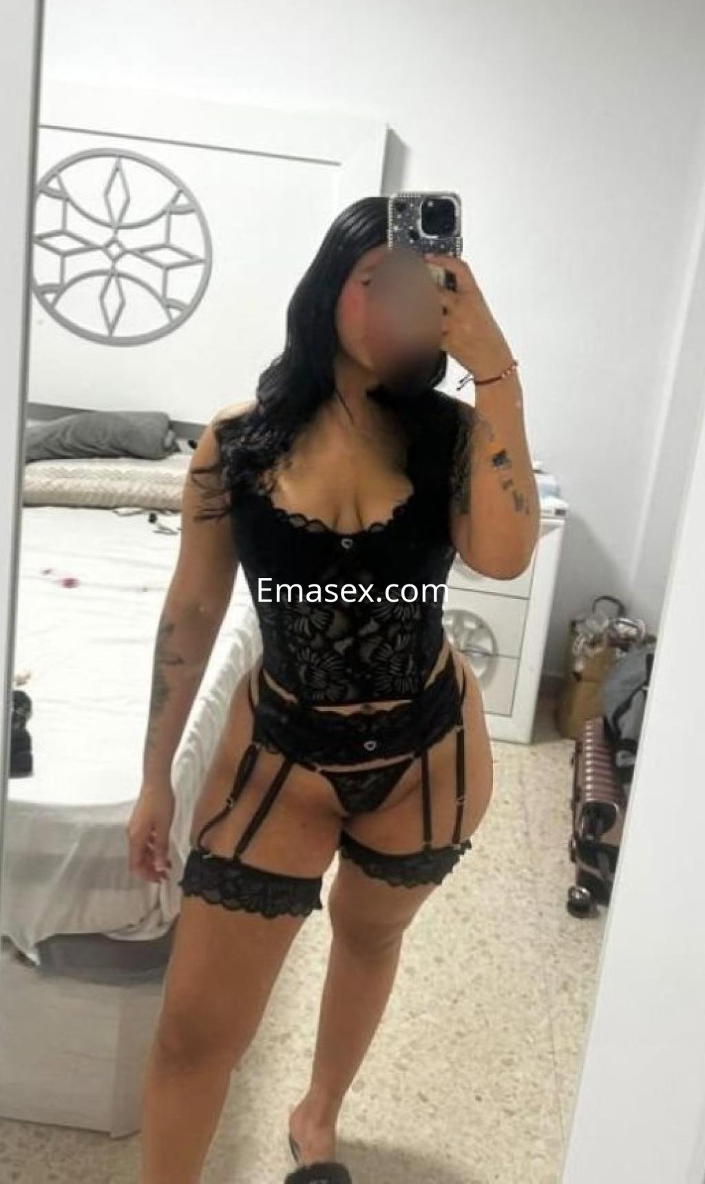 Escort en Badajoz muestra un conjunto de lencería negra con ligueros, posando frente a un espejo en un dormitorio.