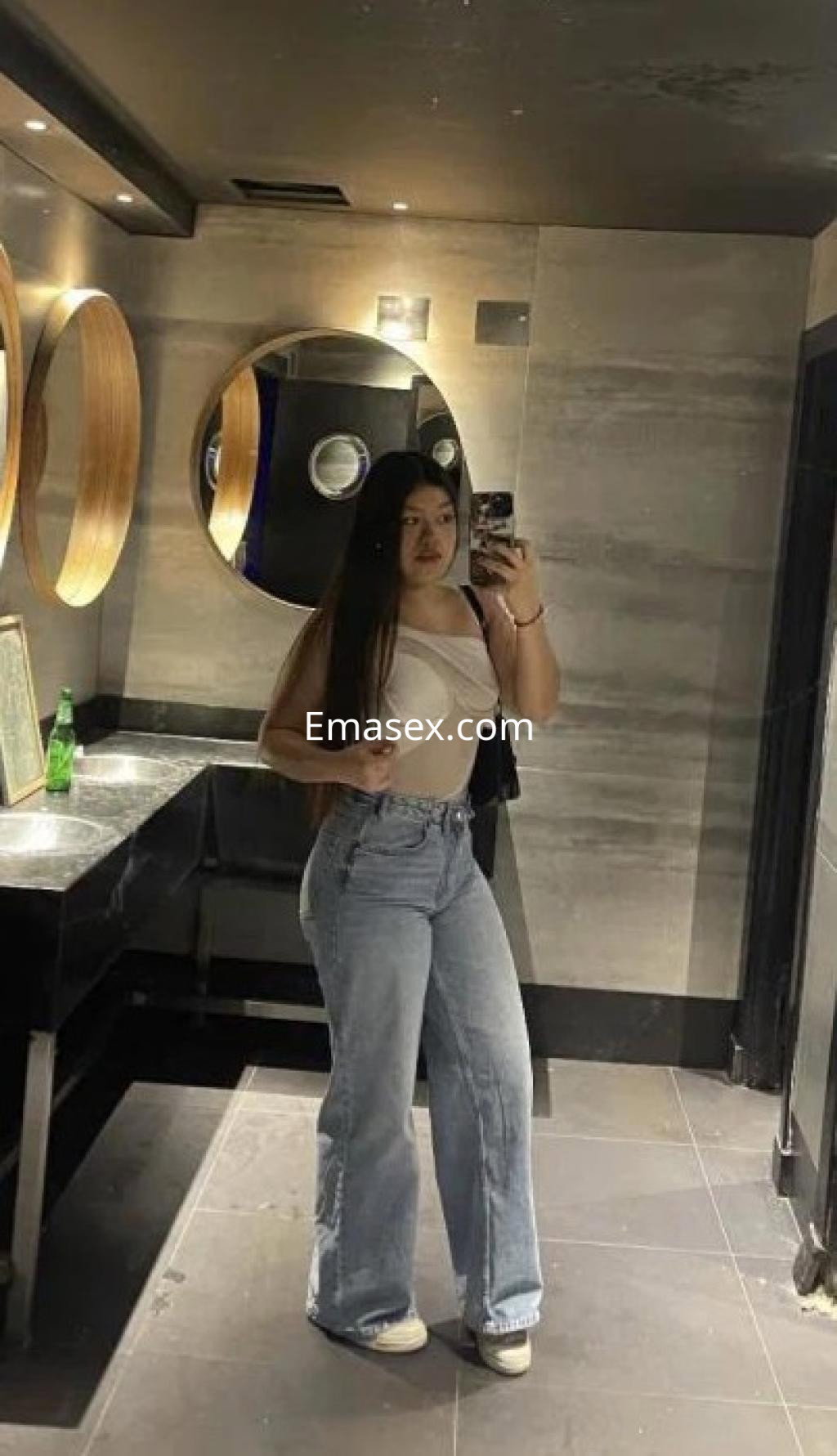 Escort en Girona, posando en un espejo con blusa clara y jeans acampanados, cabello largo y lacio.