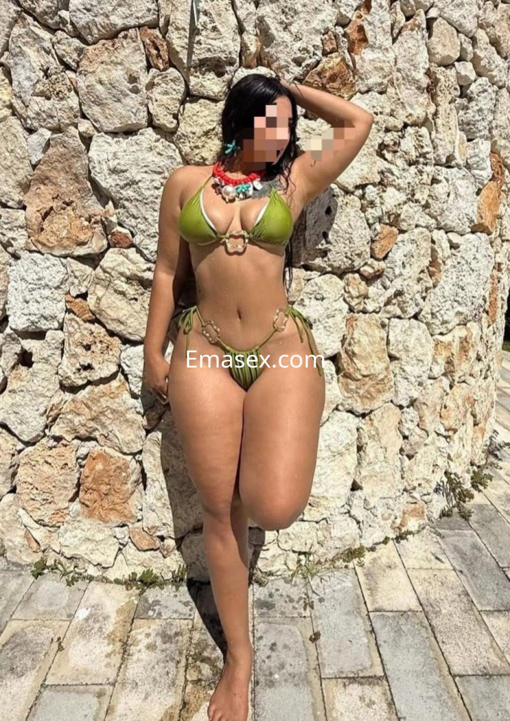 Escort en Girona apoyada en una pared de piedra, luciendo un bikini verde y con un collar colorido, en una pose coqueta.