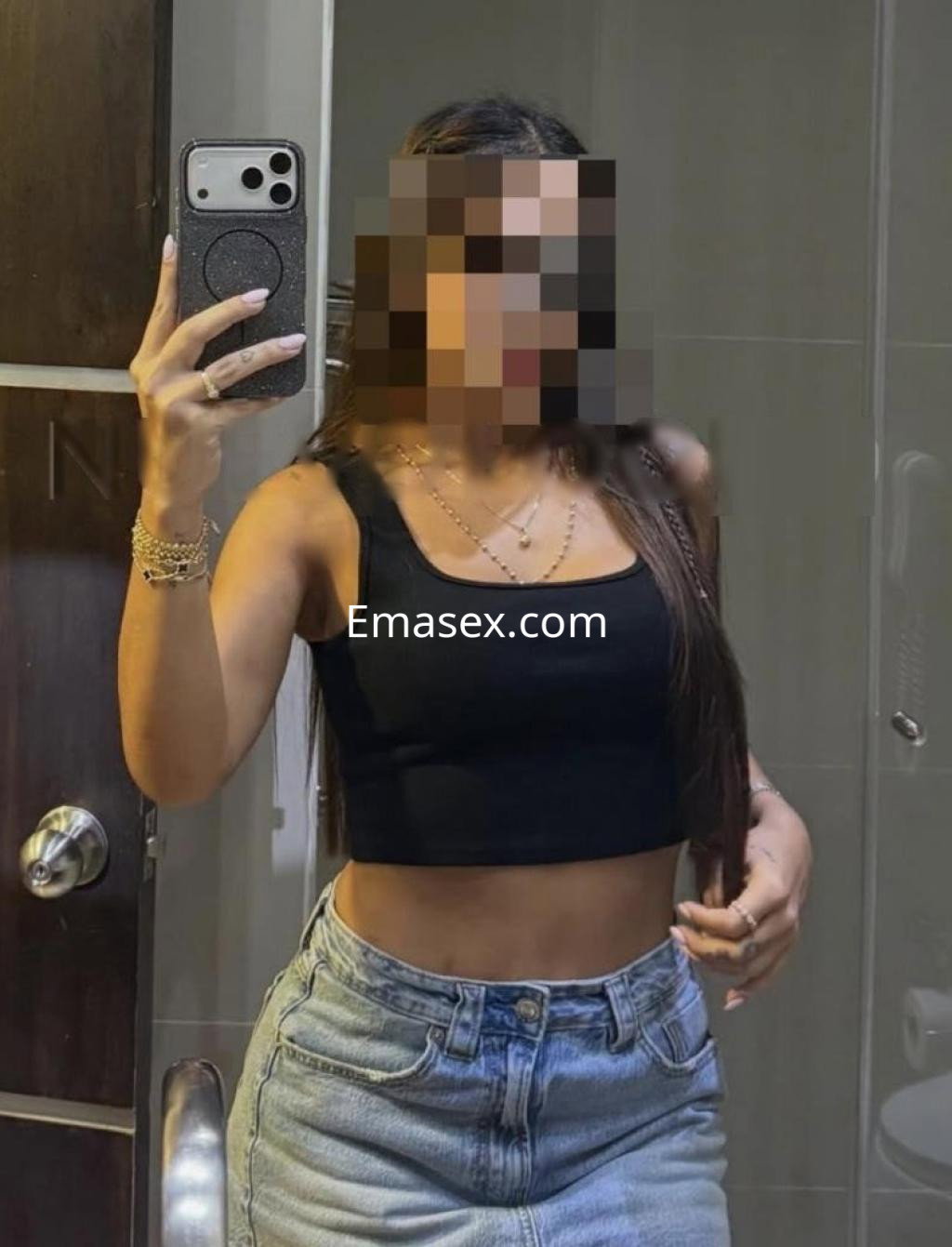 Escort en Girona se toma una selfie en un baño, vistiendo un top negro y pantalones de mezclilla, con cabello largo.