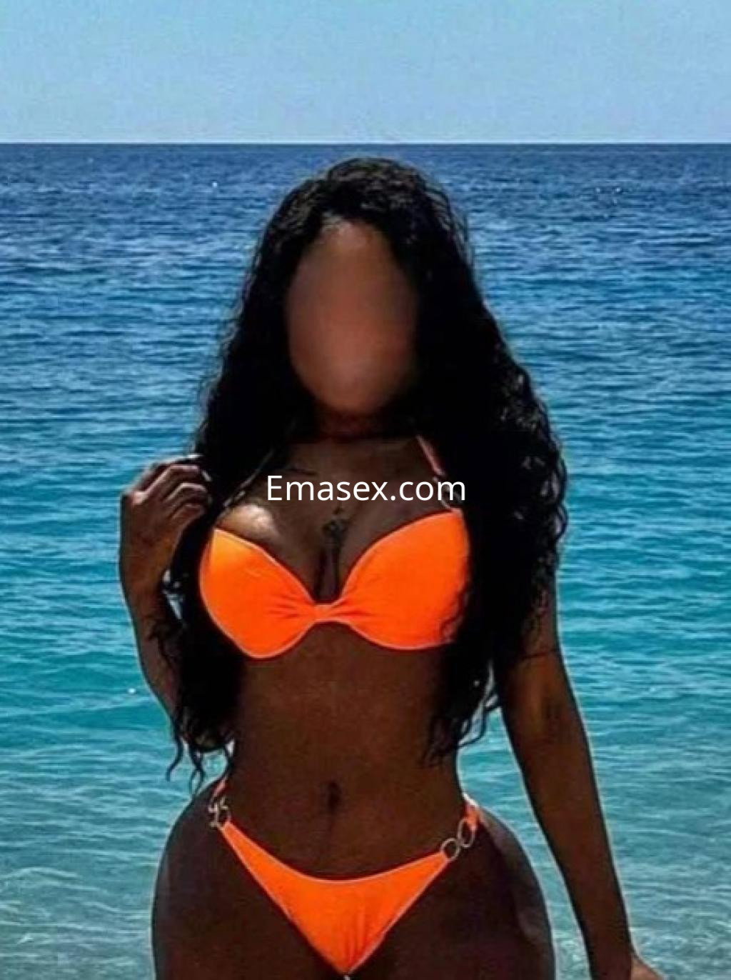 Puta en Granada con un bikini naranja, posando en la playa con el mar azul de fondo y el cabello largo y oscuro.