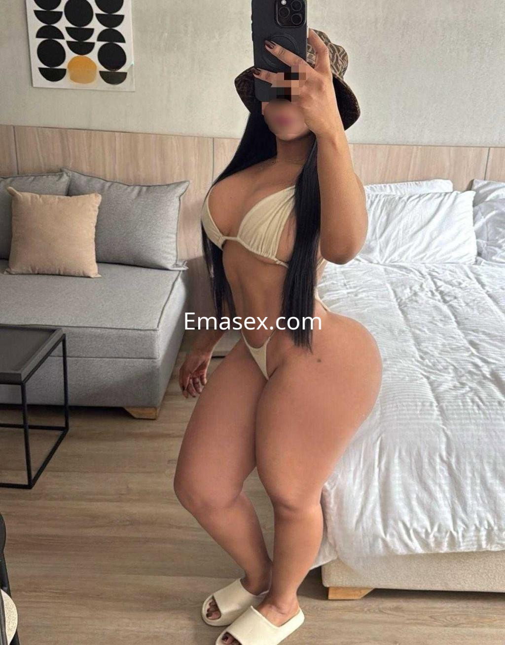 Puta con un bikini claro y sombrero, posando en una habitación moderna de Cáceres con un sofá y cama.