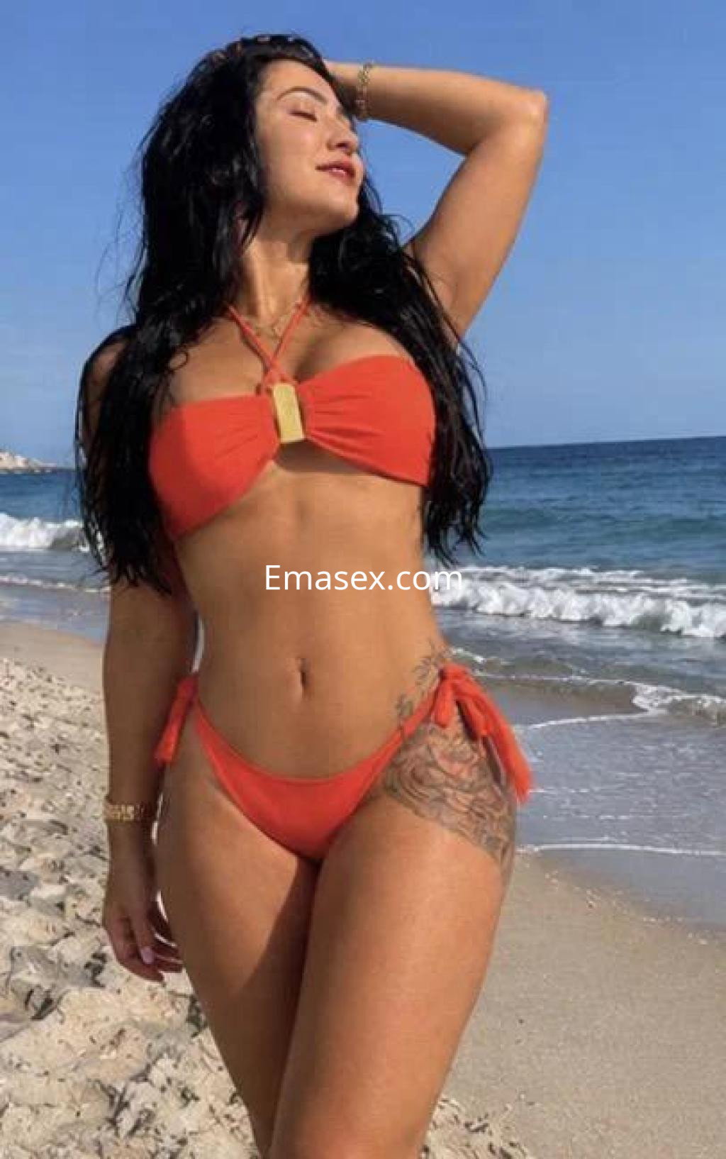Escort en Barcelona posando en la playa con un bikini rojo, cabello oscuro y una sonrisa relajada bajo el sol.