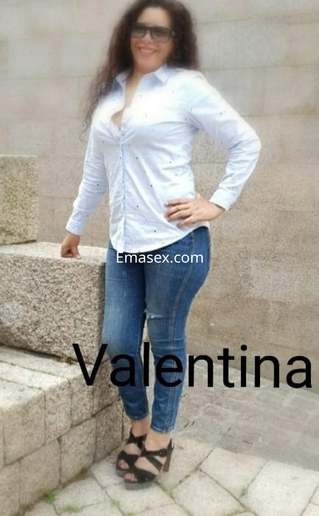 Escort en Pamplona, luciendo una camisa blanca y jeans ajustados, posando con confianza en un entorno exterior.