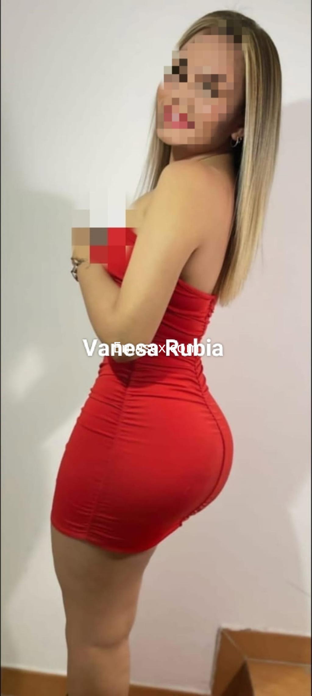 Puta en Zaragoza con cabello rubio y lacio, vistiendo un vestido rojo ajustado y posando de perfil en una habitación.