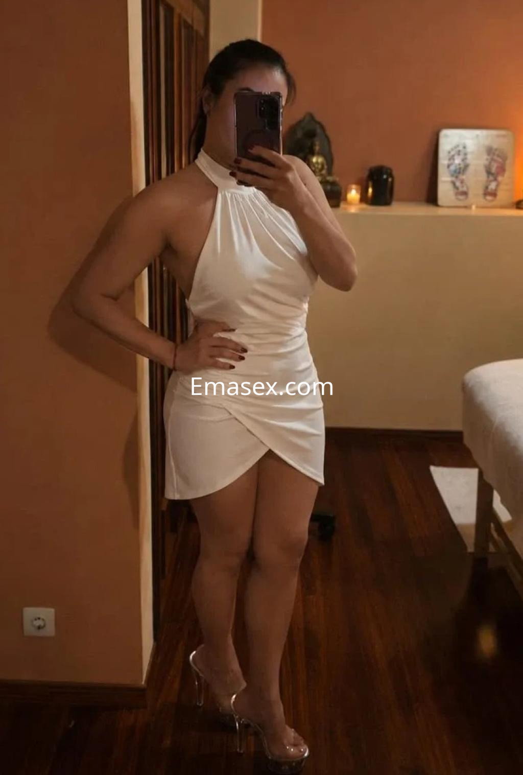 Puta en Vigo con un vestido corto blanco de tirantes y tacones transparentes, posando frente a un espejo.