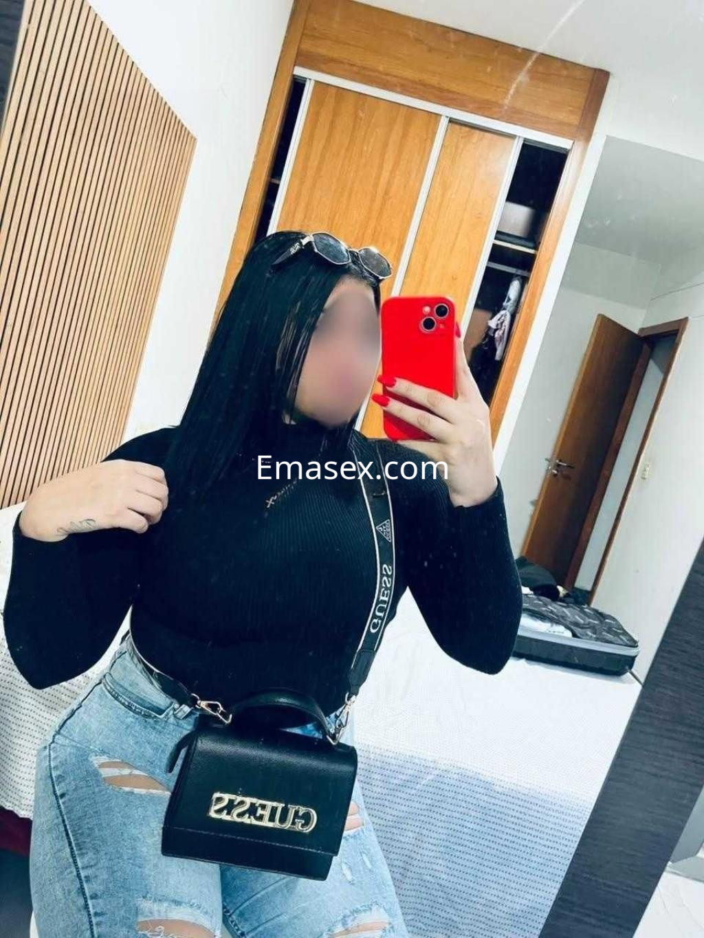Puta de Granada posando en un espejo con camiseta negra, jeans claros y bolso negro, luciendo un peinado lacio.