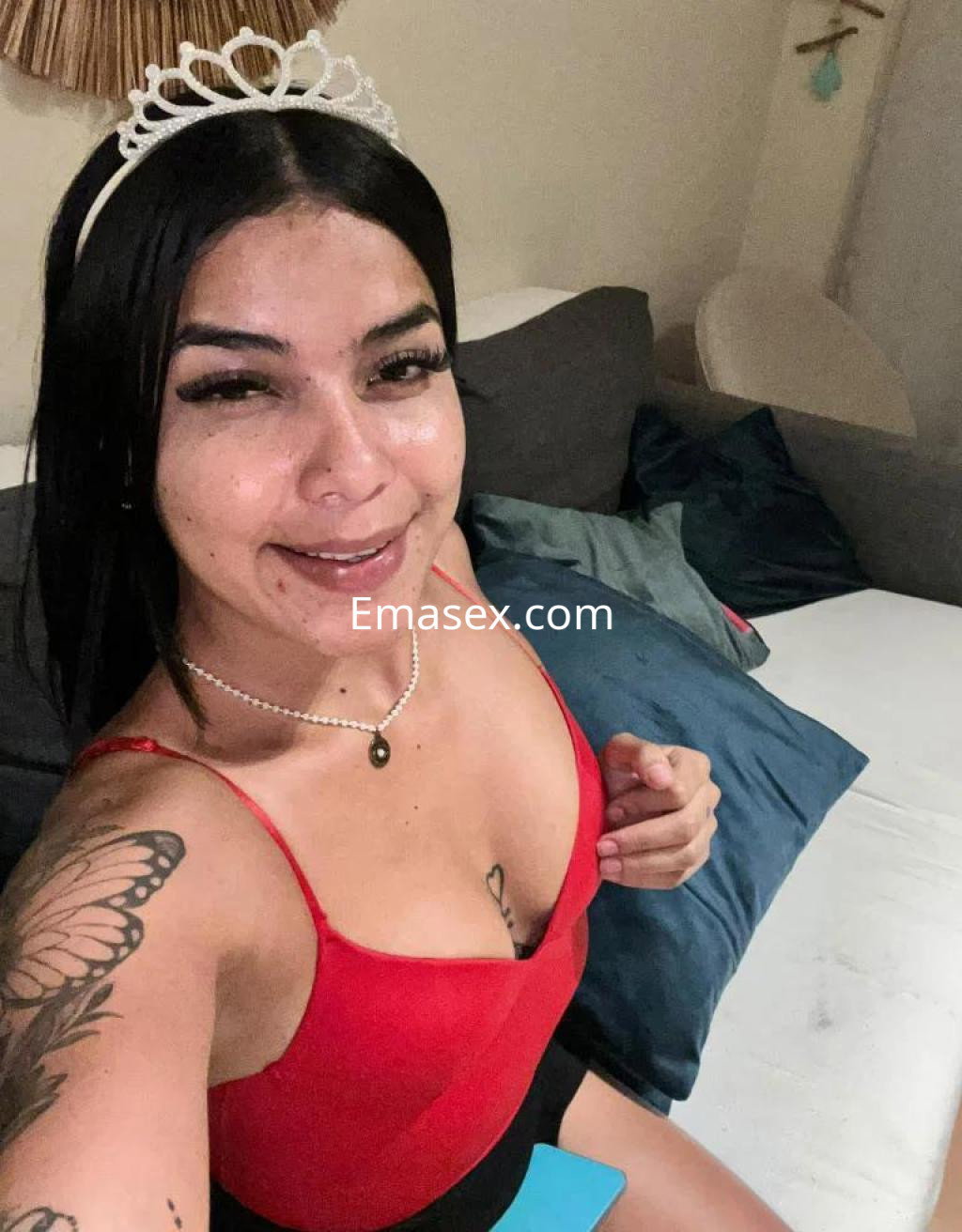 Escort en Las Palmas con cabello negro, tiara, top rojo y shorts oscuros, sonríe sentada en un sofá.