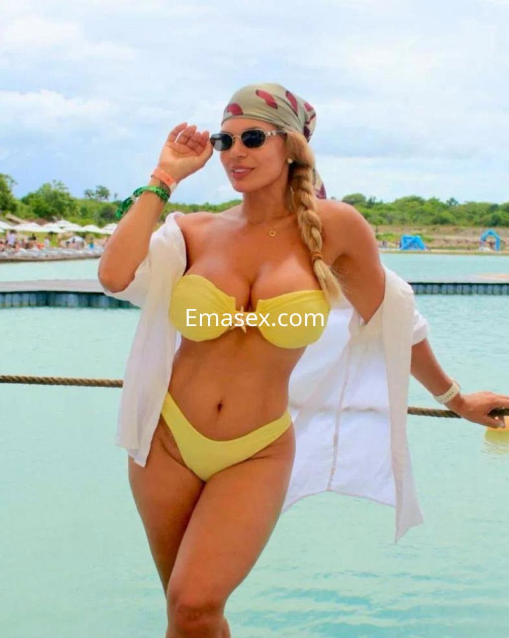 Escort en Las Palmas luce un bikini amarillo con una camisa blanca, gafas de sol y un pañuelo en la cabeza, posando junto al agua.