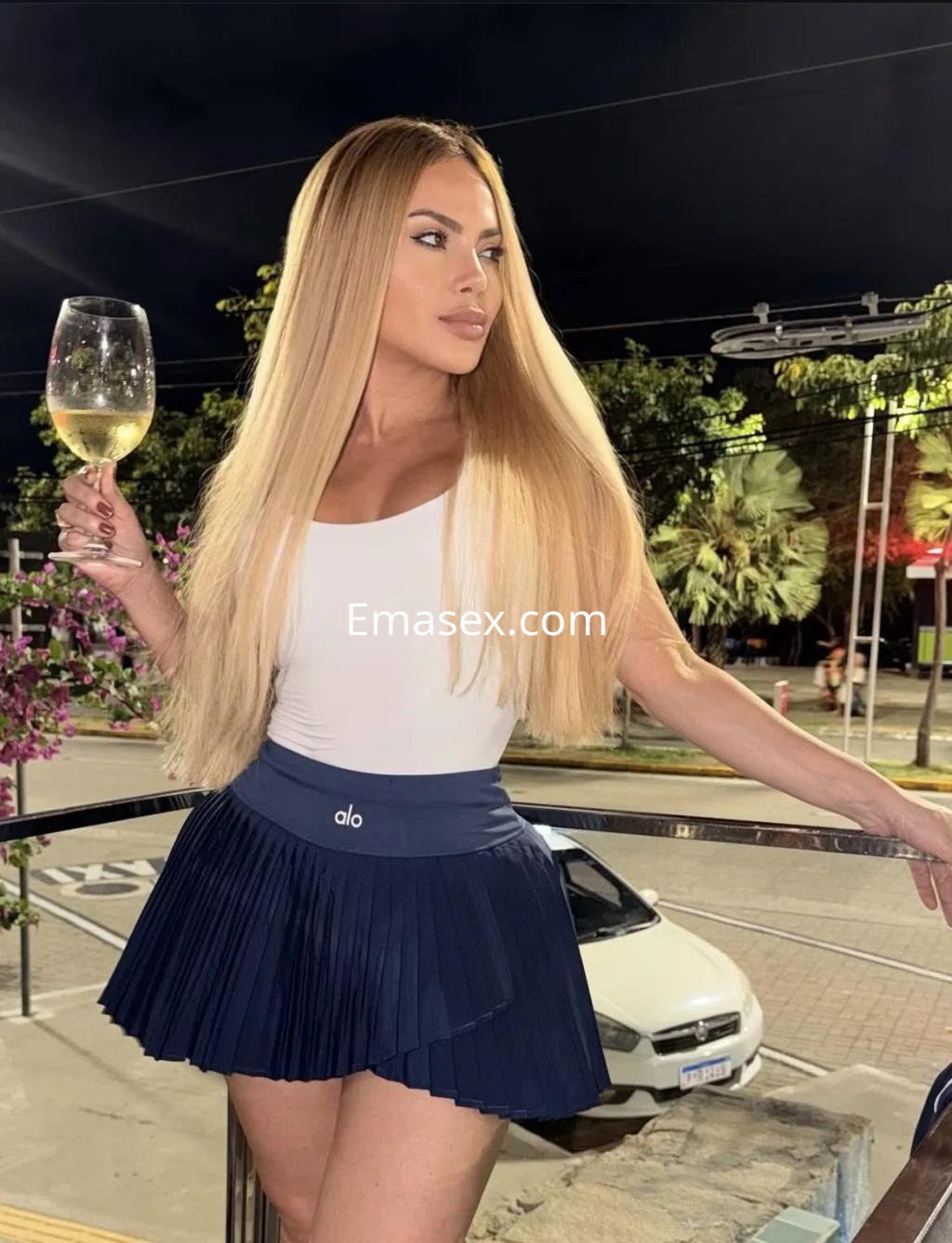 Escort en Palma de Mallorca, con cabello largo y rubio, viste una blusa blanca y una falda corta, sosteniendo una copa.