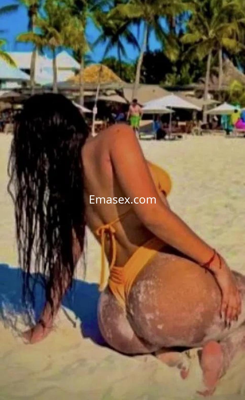 Puta en Las Palmas, con traje de baño naranja, posando de espaldas en la playa con palmeras al fondo.