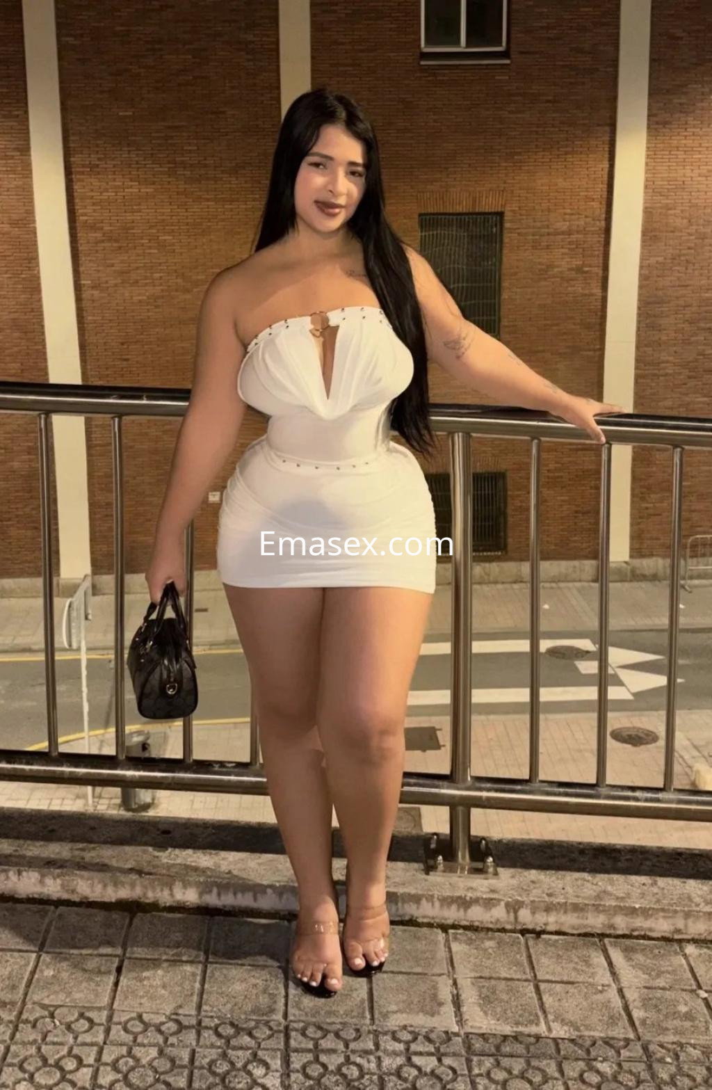 Escort en Vigo, luciendo un vestido blanco strapless con detalles en el escote, posando en una barandilla nocturna.