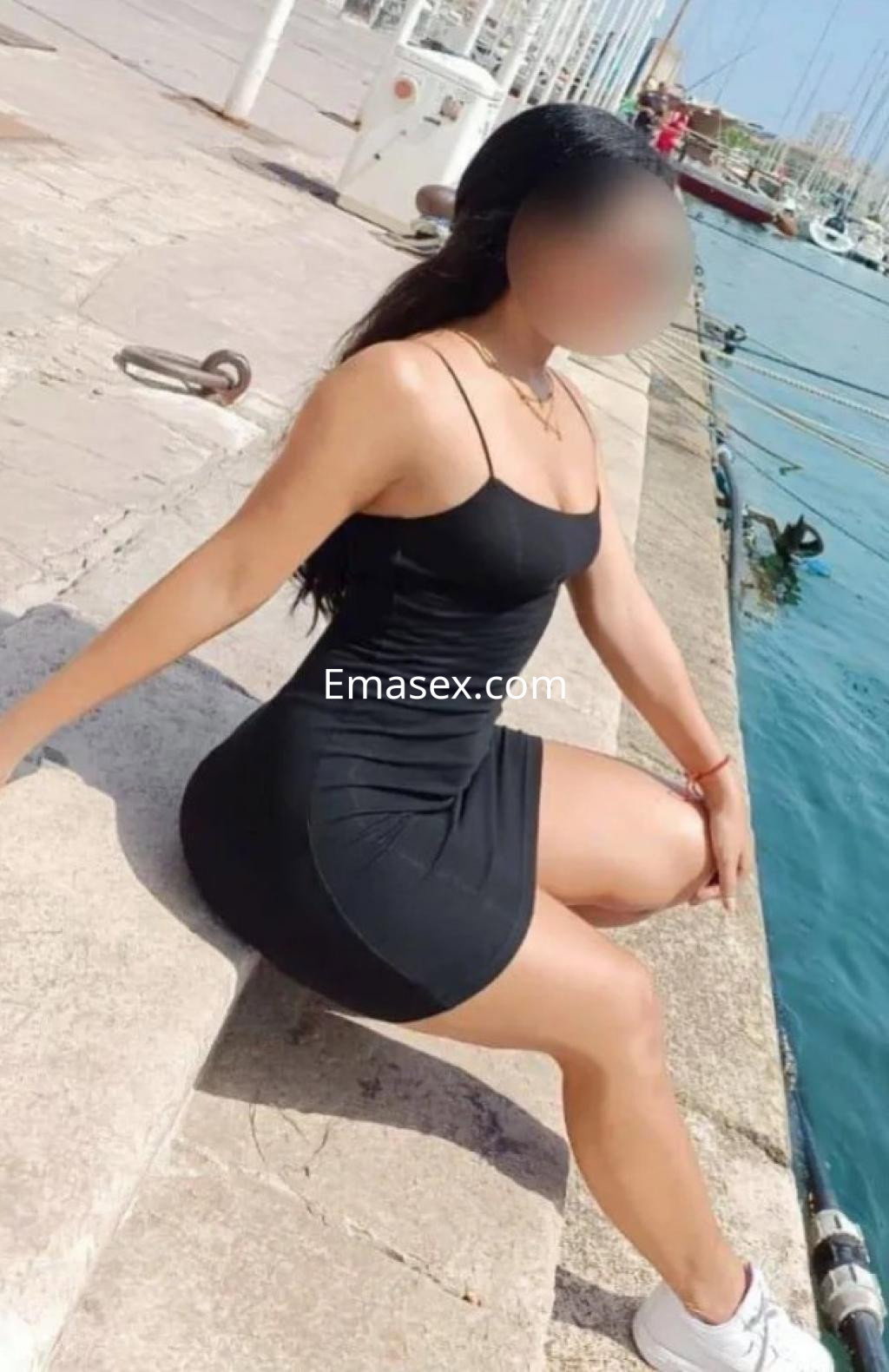 Puta en Vigo con vestido negro ajustado, sentada en un borde junto al agua, luciendo una pose relajada.