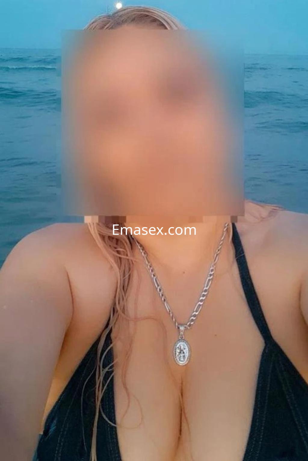 Escort en Castellón, posando en la playa con un bikini negro y un collar plateado, frente al océano al atardecer.