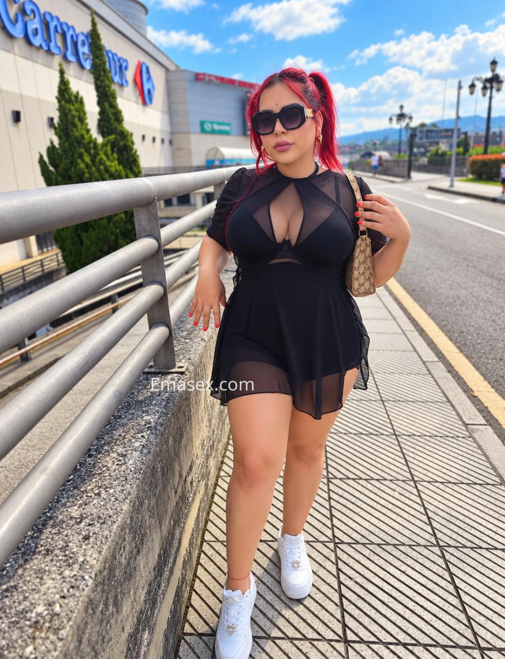 Puta trans en Alicante, luciendo un vestido transparente negro y cabello rojo, posando en una calle con tiendas.