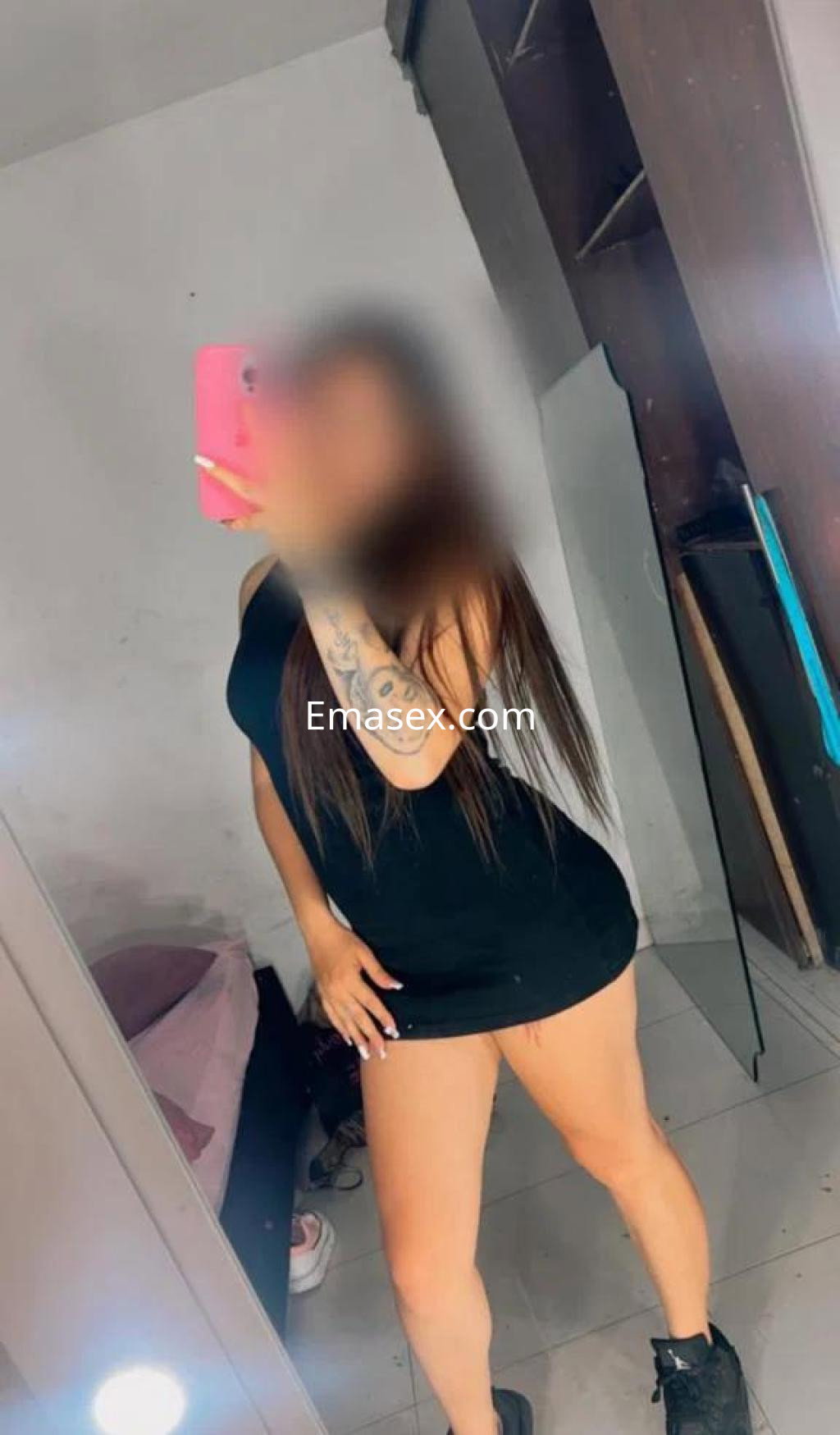 Escort en Badajoz posando frente a un espejo, con un vestido negro ajustado y tatuajes visibles en su brazo.