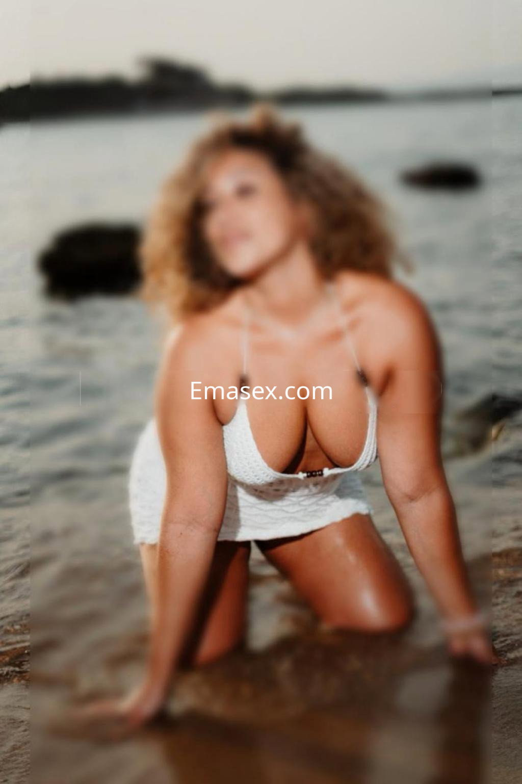 Escort en Girona con cabello rizado y vestido blanco, posando de rodillas en la orilla del mar.