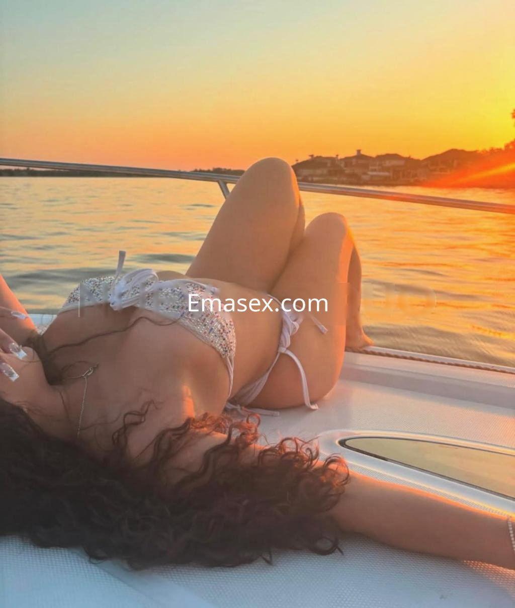 Puta en Girona, relajándose sobre un bote en el atardecer, vistiendo un bikini brillante y con cabello rizado.