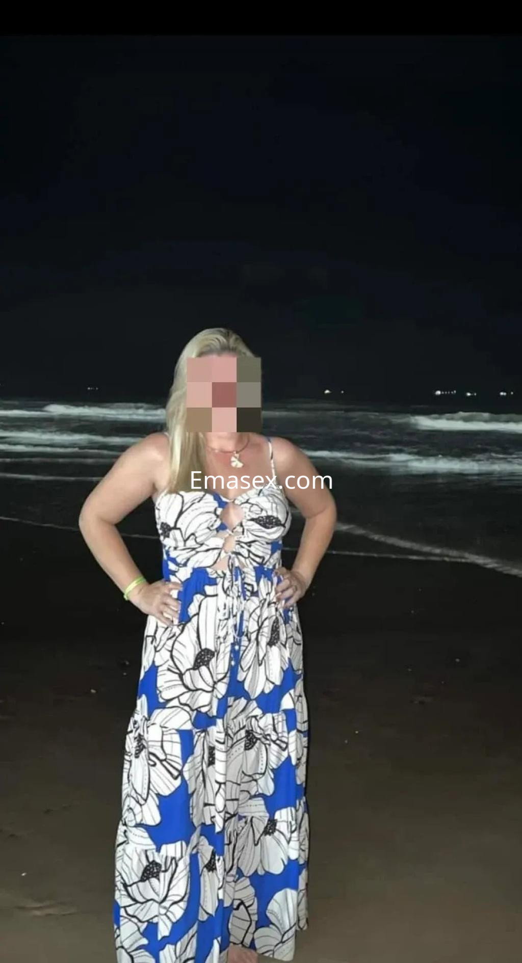 Escort en Vigo con vestido largo de flores en tonos azules y blancos, posando en la playa de noche.