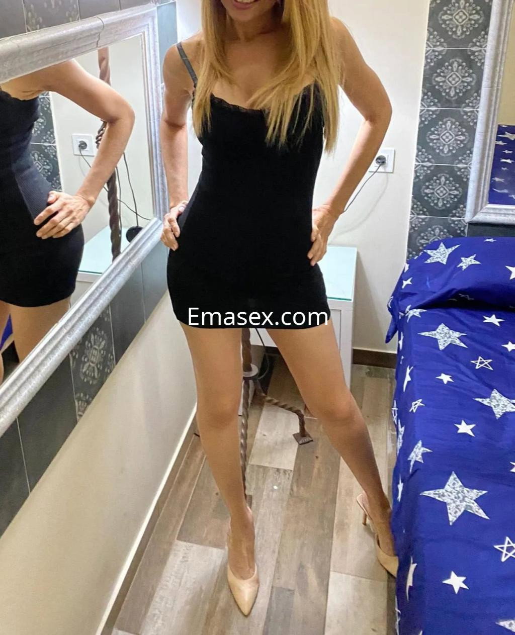 La puta en Gijón luce un vestido negro ajustado y tacones, posando con confianza en una habitación decorada.