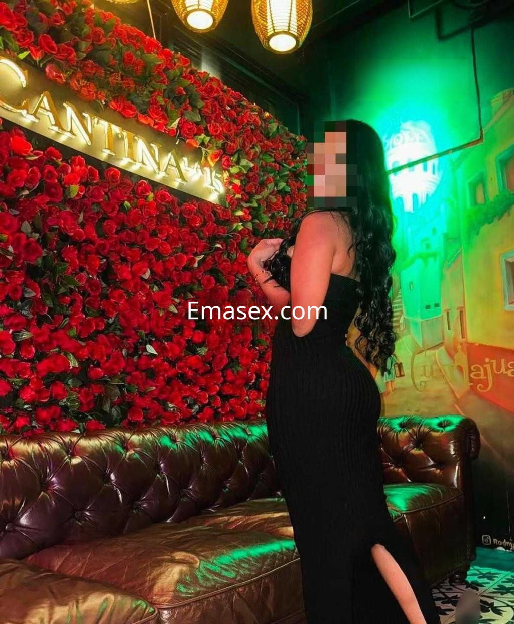 Escort en Badajoz posando con un vestido negro ajustado frente a un fondo de flores rojas.