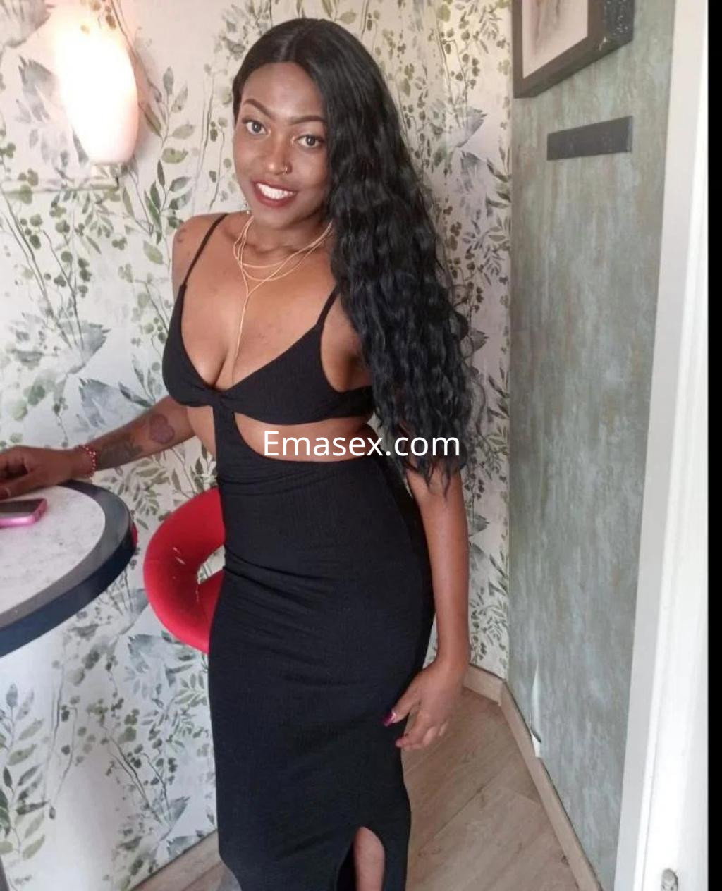 Escort en Coruña con cabello largo y ondulado, vestida con un vestido negro ajustado y abierto, posando con una sonrisa.