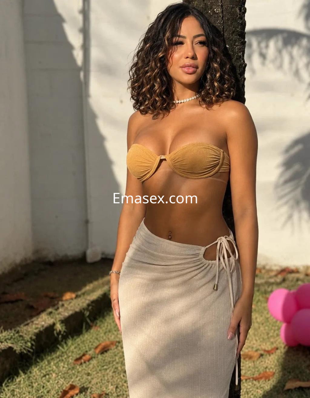 Puta en Palma de Mallorca con cabello rizado y oscuro, usando un bikini marrón y una falda larga clara, posando junto a un árbol.