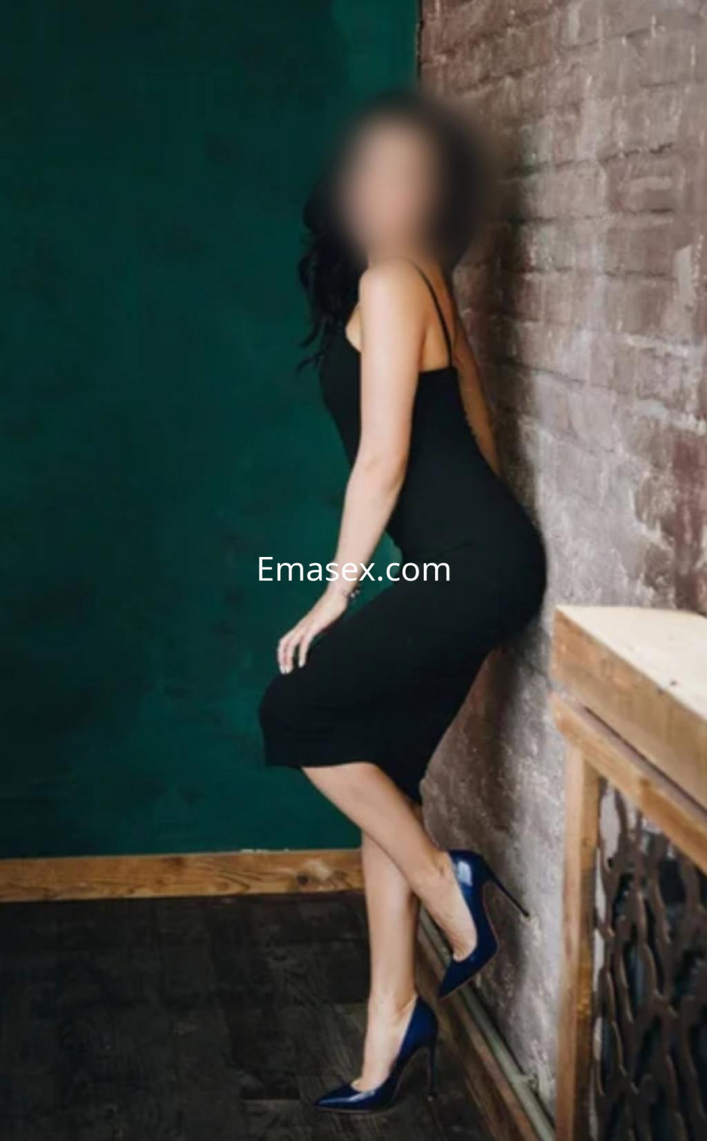 Puta posando de perfil en Santander, con un vestido negro ajustado y tacones azules, apoyada en una pared.