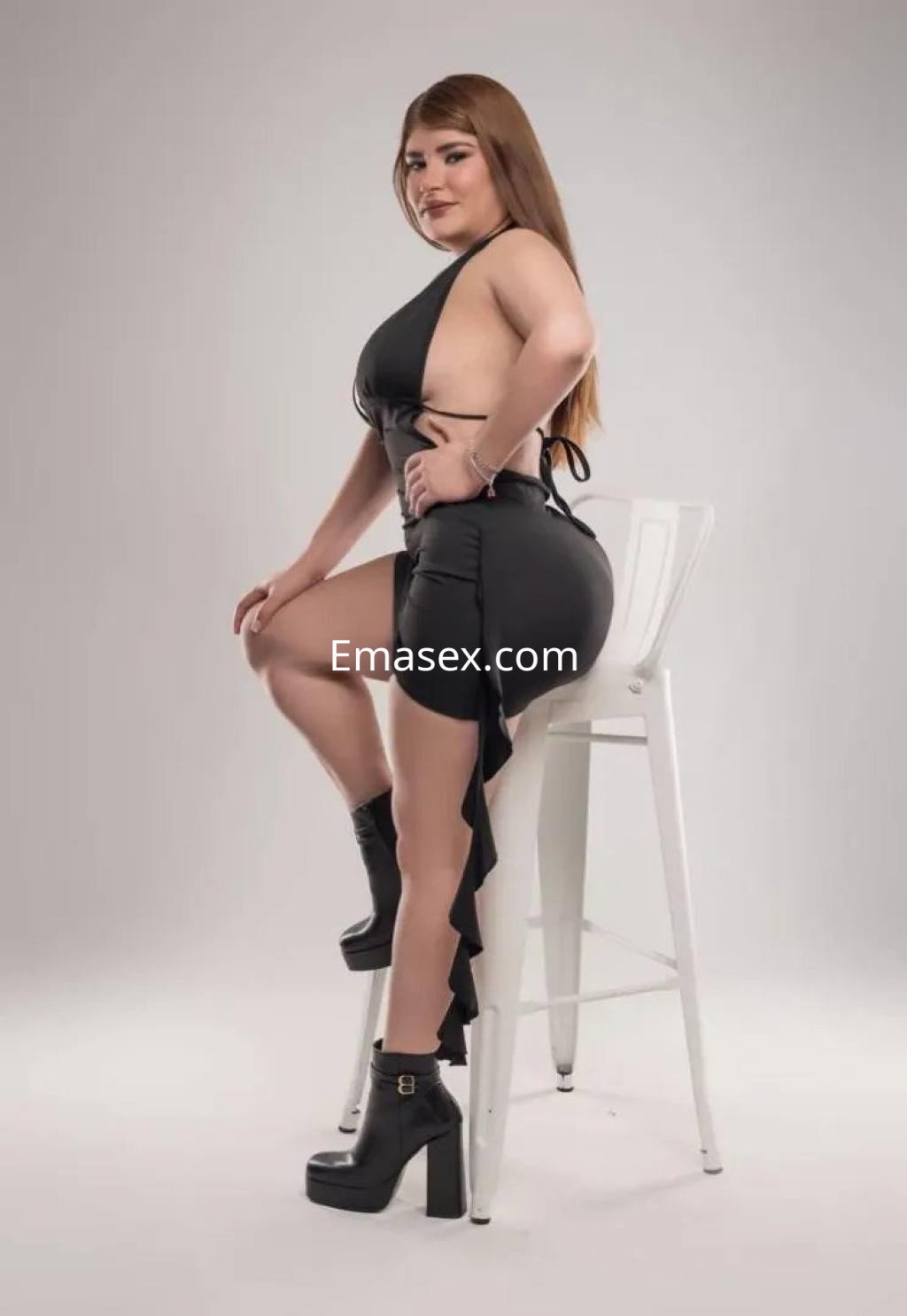 Escort en Valencia posando sentada sobre una silla, luciendo un vestido negro ajustado y botas altas.