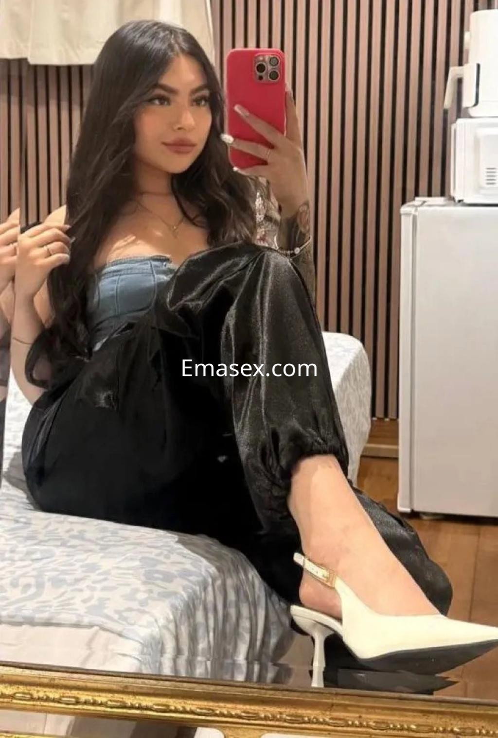 Puta posando en un sillón con pantalones negros y blusa azul, sosteniendo un teléfono en Palma de Mallorca.