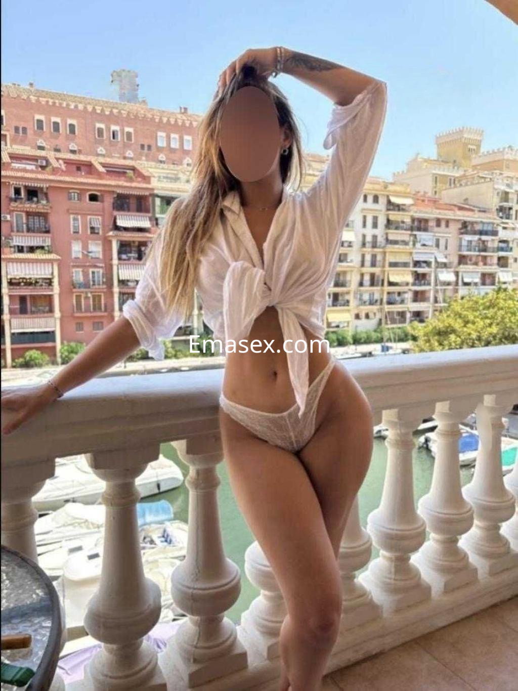 Puta en Valencia, de pie en un balcón, viste una blusa blanca anudada y lencería clara, con cabello largo y suelto.