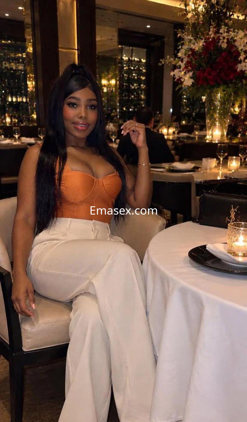 Escort en Valencia, sentada en un restaurante elegante, viste un top naranja y pantalones blancos, sonriendo.
