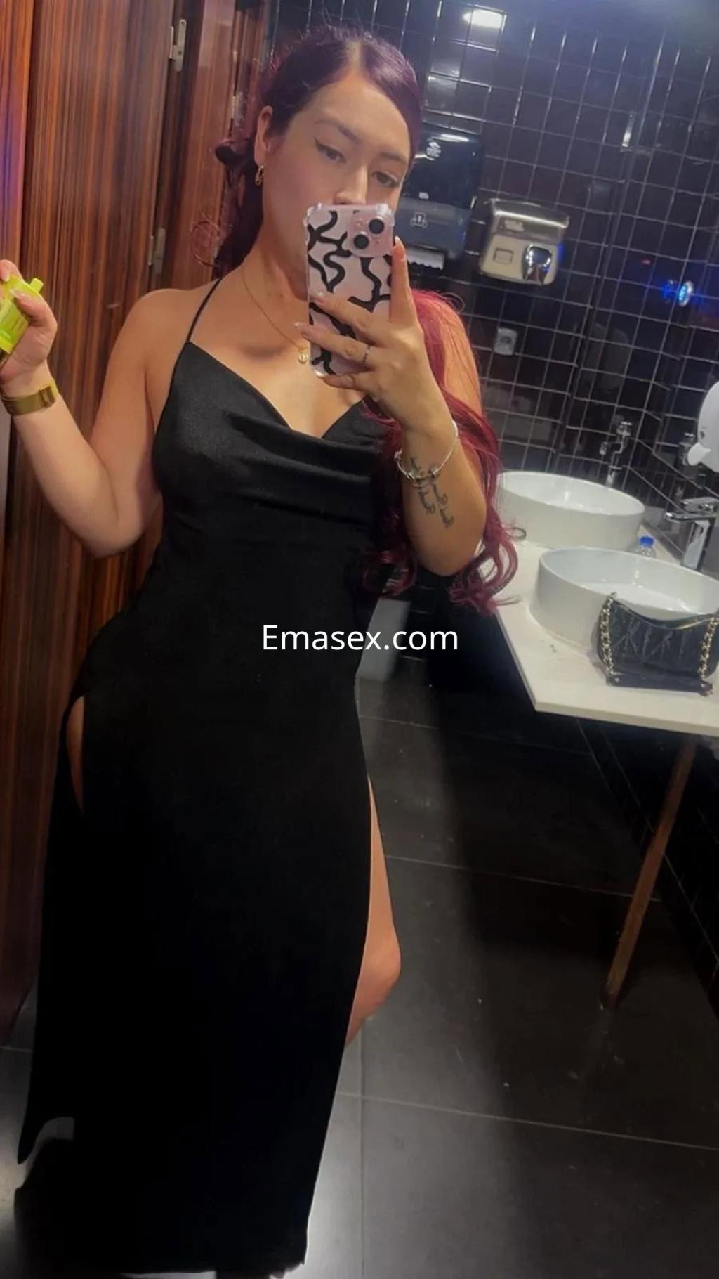 Escort de Valencia se toma una selfie en un baño, vistiendo un vestido negro con abertura y cabello largo en tonos rojizos.