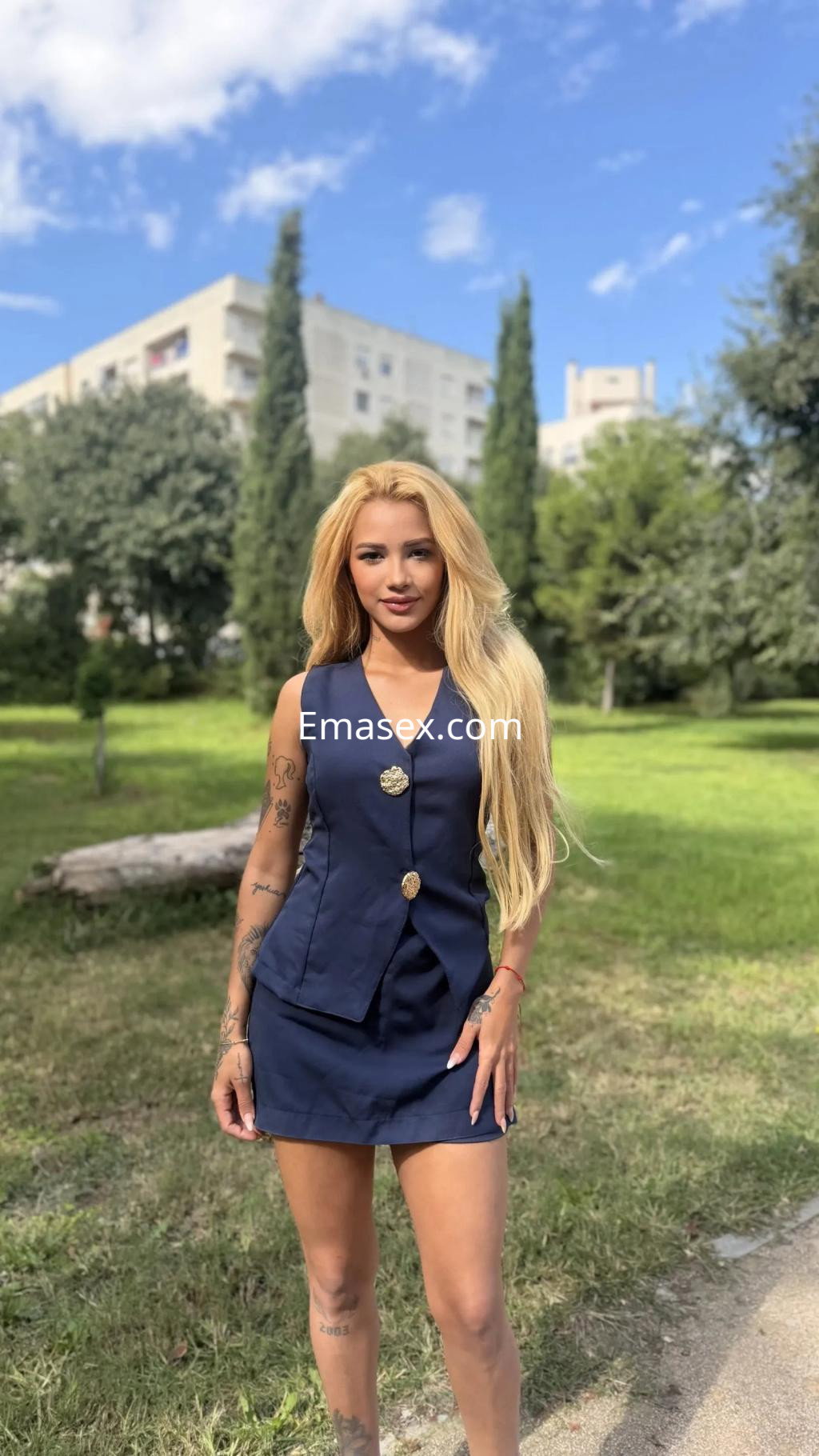 Escort en Valencia, luciendo un vestido azul oscuro y con cabello largo y rubio, posando en un parque.