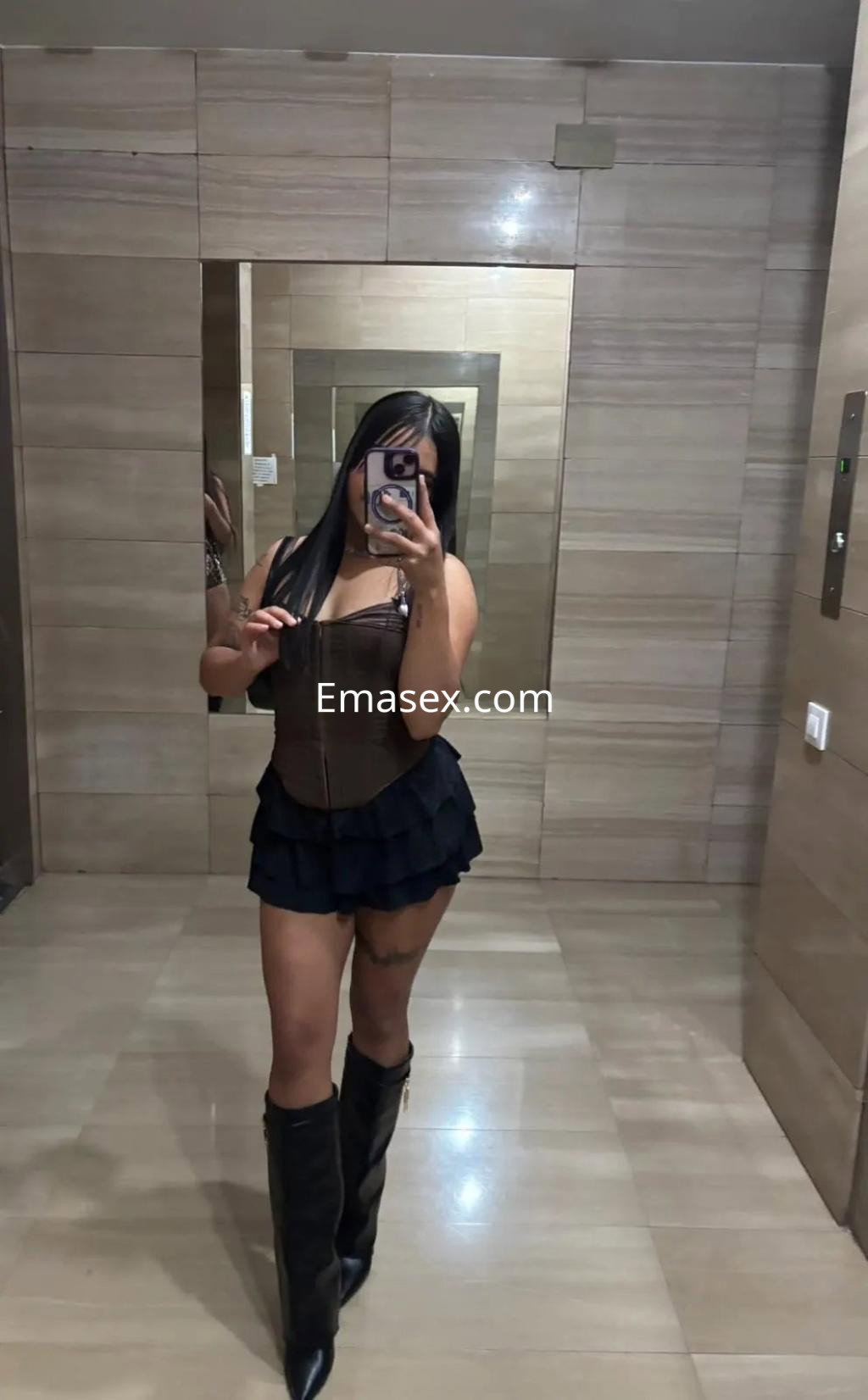 La puta en Valencia lleva un corset marrón y una falda corta, posando en un espejo con botas altas.